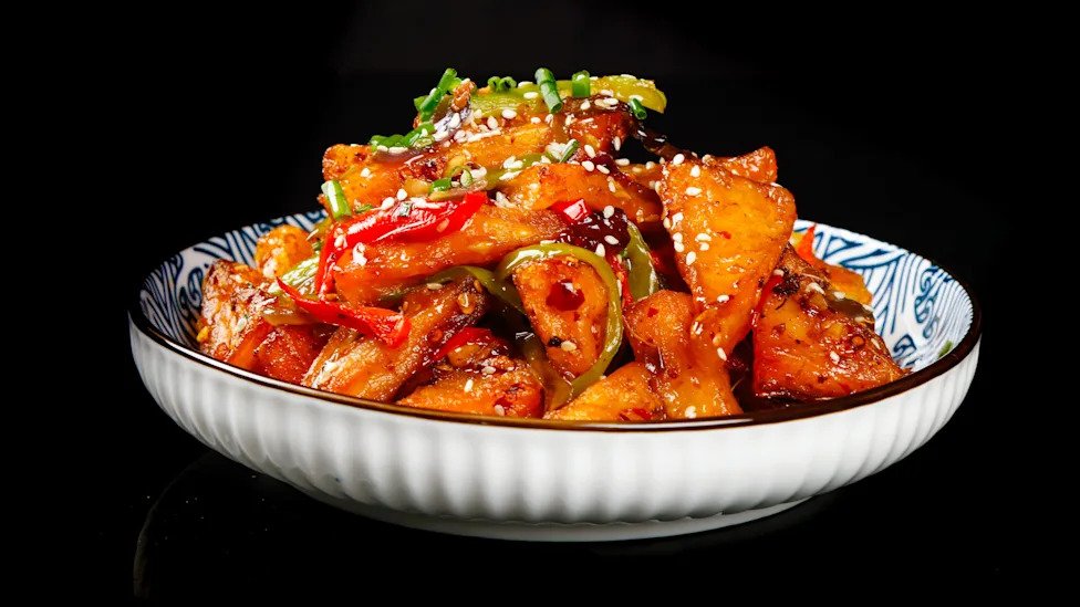 Spicy braised tofu cubes with red chili peppers, green onions and sesame seeds in traditional Chinese ceramic bowl on black background.