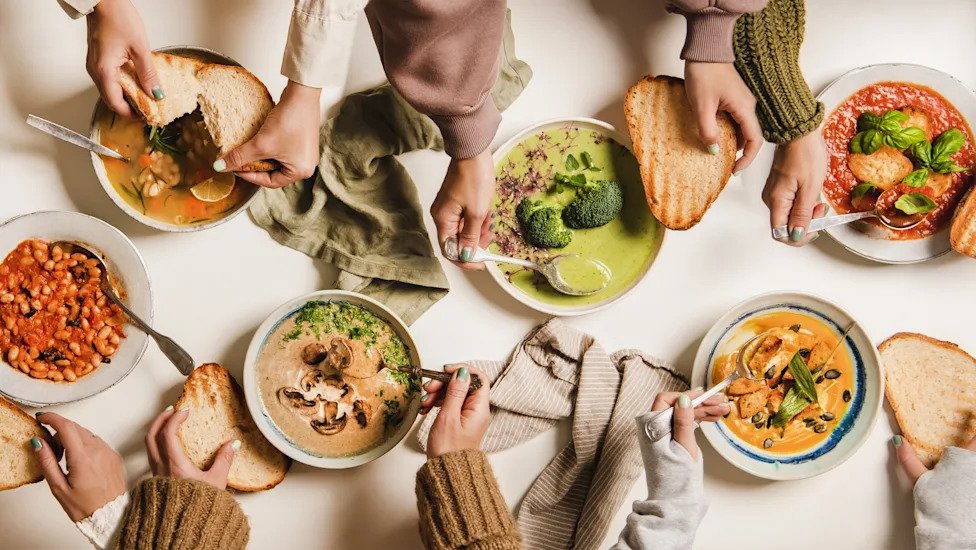 People eating Autumn and Winter creamy vegan soups. Flat-lay of peoples hands, soup plates and bread slices over plain white table background, top view. Fall and Winter food menu, vegetarian food